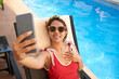 © DragonImages - Happy woman in sunglasses taking selfie for her blog when sunbathing by swimming pool