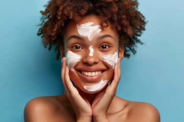 Wall Mural - Young woman with curly hair smiling using facial cream after shower standing over blue background
