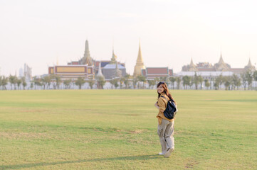 Wall Mural - Traveler asian woman in her 30s, backpack slung over her shoulder, explores Wat Pra Kaew with childlike wonder. Sunlight dances on the golden rooftops, and a carefree smile