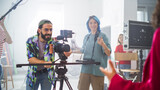 Fototapeta  - Young Caucasian Male And Female Filmmakers Collaborate On A Film Set, Adjusting Camera Equipment With Enthusiasm, Surrounded By Diverse Crew Members In A Bright, Modern Studio Environment.