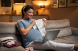 A happy pregnant woman browsing online stores for baby clothes on her laptop at home