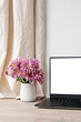 Aesthetic home office workspace, laptop computer blank screen mockup, vase with pink chrysanthemum flowers bouquet on beige wooden table, empty white wall and linen curtain background