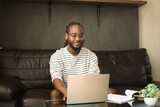 Fototapeta Nowy Jork - Pleased African man sitting on couch working with laptop or chatting in social