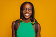 © carlesmiro - Close up portrait of a happy smiling young african american female student in green top, braids and backpack at studio isolated over yellow background.