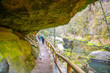 A group of hikers explores a mossy, rocky gorge with a pathway with wooden railings in Bohemian Switzerland National Park, Czechia
