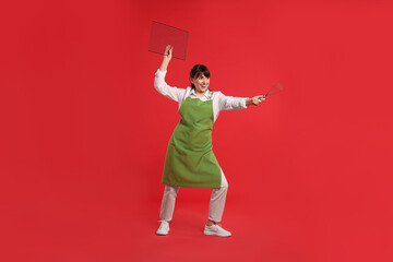 Wall Mural - Happy confectioner in apron holding professional whisk and cooling rack on red background