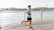 A bearded man in athletic attire runs on a sunny pier, the calm river reflecting a bustling city life in the background.