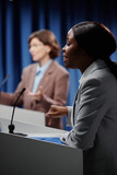 Fototapeta  - Focus on young confident female politician speaking in microphone while standing by platform in conference hall and debating with public