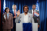 Fototapeta  - Young cheerful female politician waving hand or showing vow gesture and looking at camera while standing by platform against delegates