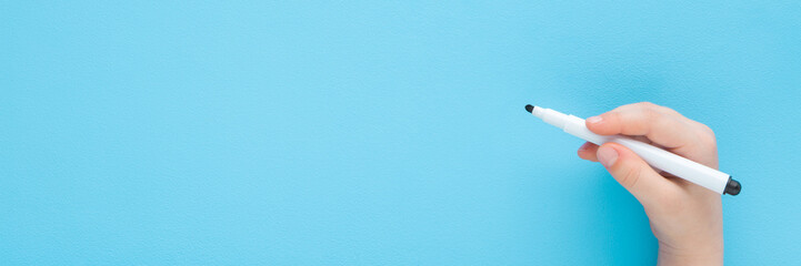 Baby boy hand holding black white color pen and writing on light blue table background. Pastel color. Closeup. Wide banner. Empty place for text. Top down view.