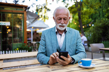 Mature guy is connected on a smartphone in the bar. Happy senior man using smartphone app while sitting in restaurant terrace and drinking coffee. Businessman using phone with new trend technology.