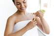 Close-up of a pretty hispanic or brazilian young woman, wrapped in a white towel, applying moisturizing serum on her hands after shower, smiling. Caring of a body and hands