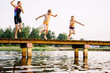 Teen girl in swimsuit and two smaller boys jumping from a bridge into a lake at sunset.