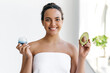 Beautiful charming well-groomed brazilian or hispanic young woman wrapped in a towel stands on white background, holds an avocado and moisturizing cream in her hands, smile at camera. Skincare concept