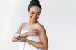 Caring of a body. Happy beautiful hispanic or brazilian young woman standing on a white background wrapped in white towel, gently applying moisturizing cream on body, smiling