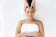 Top-view on facial massage. Relaxed brazilian or hispanic woman lies on a couch in a salon during a forehead massage. Female massage therapist doing general forehead massage to a cute girl