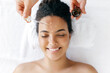 Top view of a gorgeous well-groomed smiling hispanic or brazilian woman, wrapped in a white towel, lying during a procedure with a cosmetologist which applying on her forehead hyaluronic serum