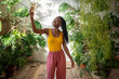 Joyful African American woman blogger takes selfies surround grown indoor tropical plants for blog. Happy black girl plants lover smiling make self photo on mobile phone in home garden urban jungle.
