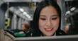 Cute Chinese woman sitting on chair in public transport. Using smartphone. Beginning to laugh. Continuing watching video or images on Internet. Texting with friends or family member.