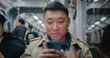 Camera view of Asian man sitting next to window in public transport. Using smartphone while listening to music in earphones. Checking weather or stop outside. Tapping on screen with both thumbs.