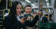 Asian girl standing in middle of large wagon of train or metro. Watching videos or images on smartphone. Man besides peeks why woman laughing. Also beginning smiling. Having fun together.
