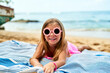 Smiling girl with pink sunglasses lies on blue striped towel at sunny beach. Happy child enjoys summer, sand, sea vacation. Playful kid in swimwear relaxes by ocean, fun holiday travel with family.