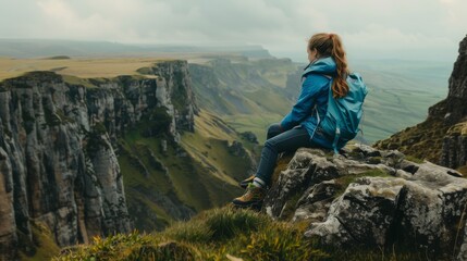 A side view of an anonymous woman in a blue jacket and boots sitting on the edge of a rocky cliff, observing the scenic view of a green mountainous valley.