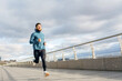 A spirited runner in a light blue jacket and black pants strides confidently along an expansive river walkway.