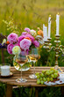 Romantic sunset wedding table decor for a couple with vintage chandelier, burning candles, snacks, white wine, fruits, ranunculi flowers. Summer vibes, field or meadow, outdoors event