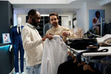 Fototapeta Tulipany - Fashion boutique customer getting assistance from employee while choosing shirt size. African american man holding apparel on hanger and discussion formal outfit with showroom worker