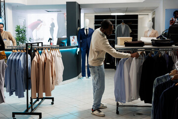 Wall Mural - African american man searching shirt size while choosing formal wear outfit in clothing store. Boutique customer browsing rack with hanging apparel in shopping mall outlet