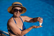 Portrait of an old woman in a straw hat, sunglasses and a swimsuit applying sunscreen to her skin while relaxing by the pool. 