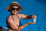 Fototapeta Zwierzęta - Portrait of an old woman in a straw hat, sunglasses and a swimsuit applying sunscreen to her skin while relaxing by the pool. 