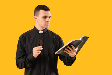 Wall Mural - Young priest with Holy Bible and cross on yellow background