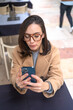 Serious middle-aged woman using smartphone sitting outdoor cafe. Focused businesswoman in glasses dressed trench coat holding smartphone in hand looking screen. Vertical photo high angle view