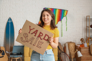 Canvas Print - Young woman holding cardboard with text YARD SALE in room of unwanted stuff