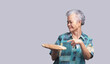An elderly Asian woman holding and pointing to a wooden tray with a smile while standing on a gray background.