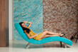 Good-looking young woman make-up and long hair wearing yellow dress posing sitting on blue trendy comfortable sofa against white wall background. Fashion and interior design concept