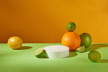 Empty round podium on green table decorated by orange, limes, apple and lemon against on orange background at front view. Cooking or drinking template with copy space