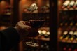 Close-up of a hand gently holding a red wine glass, set against a backdrop of a warmly illuminated wine cellar filled with numerous bottles.