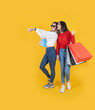 Full body young two caucasian friends woman wear casual jean and shirt hold carry hand paper bags packages showing pointing aside copy space. Yellow studio background. Copy space. Sale buy concept.