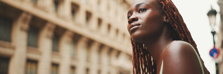 Wall Mural - Gorgeous woman with African braids wearing top stands on the sidewalk next to the road waiting for taxi, Panorama