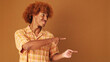 Stylish curly guy points to the side at pointing fingers to show mockup space, isolated on brown background in the studio