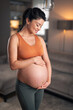 A joyful pregnant woman holding and gazing at her stomach while standing in the living room
