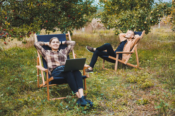 Wall Mural - Concentrated woman in the garden working on a laptop while sitting on a chair among the trees. A woman works remotely from the garden, thinking about a new business or creative idea.