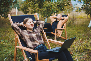 Wall Mural - Concentrated woman in the garden working on a laptop while sitting on a chair among the trees. A woman works remotely from the garden, thinking about a new business or creative idea.