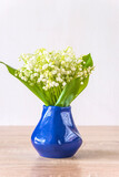 Fototapeta Krajobraz - Happy May Day greeting card; bouquet of lilies of the valley in blue vase on a wooden table and white background; copy space; vertical picture