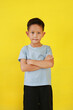 Portrait of smiling Asian boy age about 7 years old standing and cross arms over chest looking straight at camera isolated on yellow studio background.