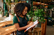 A beautiful woman in a green t-shirt and an apron is writing on a clipboard and standing in a plant shop.