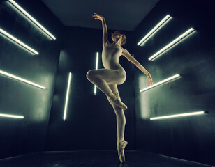 ballerina in a bodysuit and tights poses stretching in a dark photo studio in the light of lamps